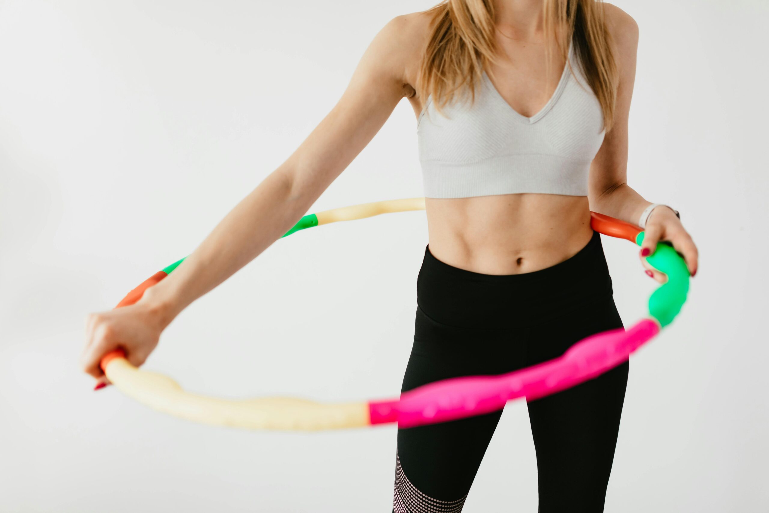 Crop faceless fit lady in black leggings and sports bra practicing fitness exercises with hula hoop against gray background